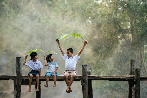 아시아 소년과 어린 소녀들은 나무 다리에 앉아 머리에 바나나 잎을 가지고 노는 것을 즐거워하고 함께 웃으면서 웃고, 복사 공간, 시골 풍경 스타일 개념