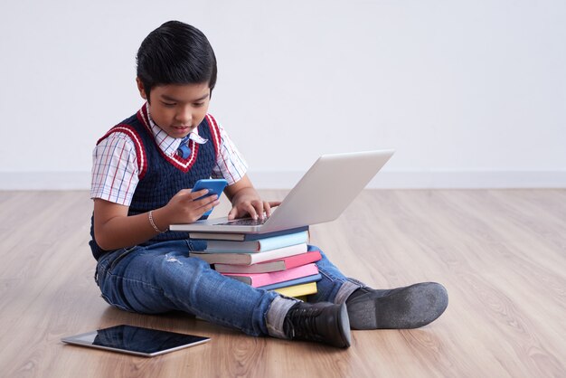 쌓인 책에 태블릿 및 노트북 바닥에 앉아 스마트 폰을 사용하는 아시아 소년