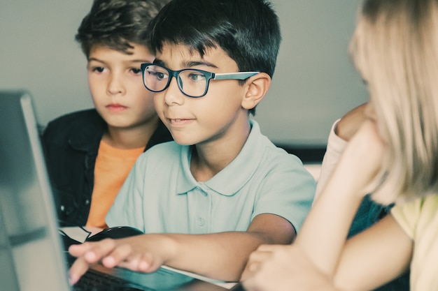 Asian boy typing on laptop keyboard and classmates sitting at table, watching him and doing task together