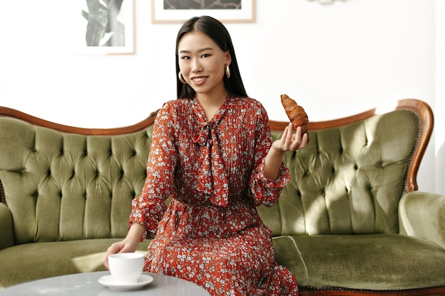 Asian brunette attractive woman in stylish red floral dress smiles sincerely, sits on soft green sofa, takes cup of tea and holds croissant