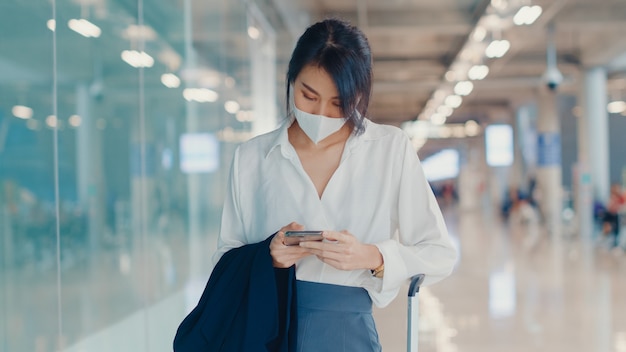 공항에서 국내선 터미널에 수하물과 함께 걷는 체크 탑승권 스마트 폰을 사용하는 아시아 비즈니스 소녀.