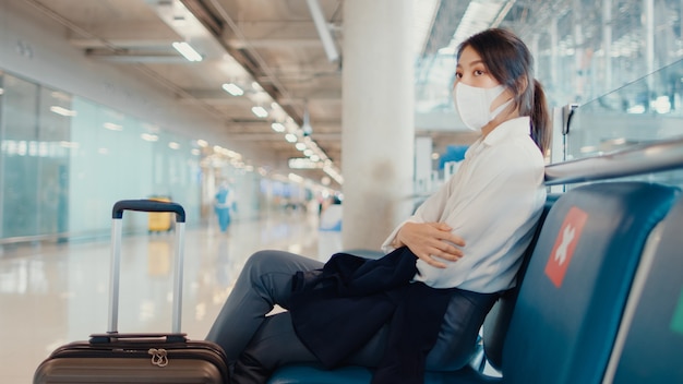 Азиатская бизнес-девушка с багажом, сидя на скамейке, ждет и ищет партнера для полета в аэропорту.