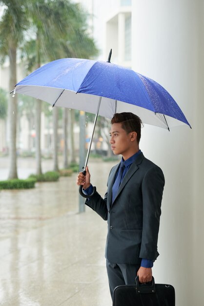 雨の中に傘が付いている通りに立っているアジア系のビジネスマン