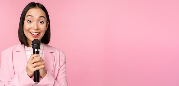 Азиатская деловая женщина произносит речь, держа микрофон и улыбаясь, стоя в костюме на розовом фоне