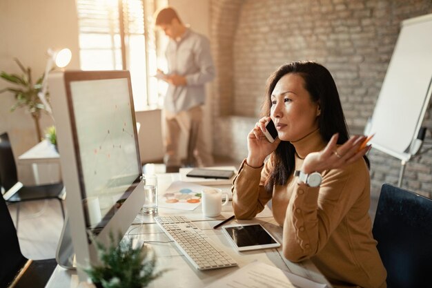 Asian businesswoman reading charts on desktop PC and communicating on cell phone in the office