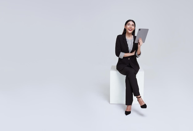 Азиатская деловая женщина улыбается в строгом костюме, сидя на стуле с помощью планшета и глядя в копировальное пространство