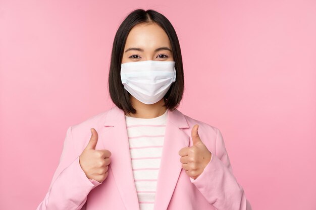 Asian businesswoman in suit and medical face mask showing thumbs up recommending wearing personal protective equipment in office during covid19 pandemic pink background