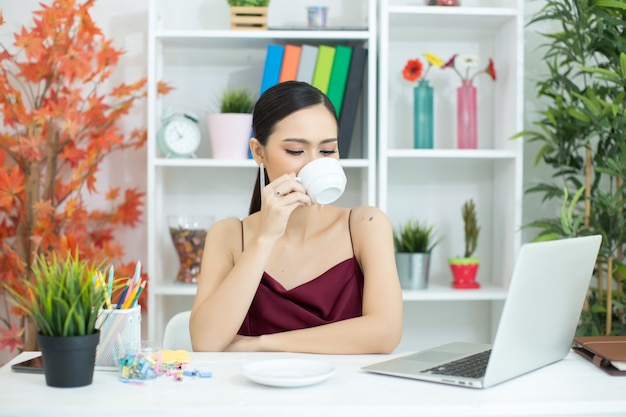 Азиатская коммерсантка принимает перерыв на чашку кофе после работы на портативном компьютере на столе