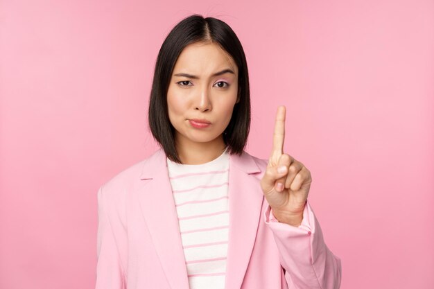 Asian businesswoman with serious concerned face expression showing stop motion taboo prohibit gesture disapprove smth bad standing over pink background