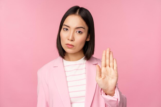 Asian businesswoman with serious concerned face expression showing stop motion taboo prohibit gesture disapprove smth bad standing over pink background