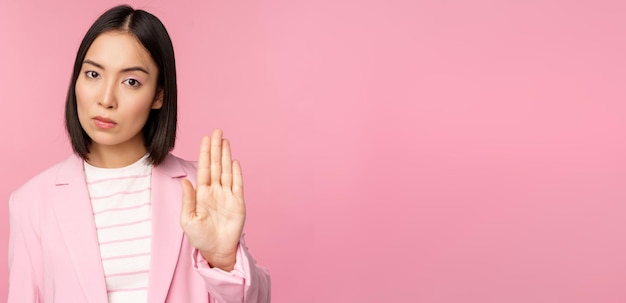 Asian businesswoman with serious concerned face expression showing stop motion taboo prohibit gesture disapprove smth bad standing over pink background