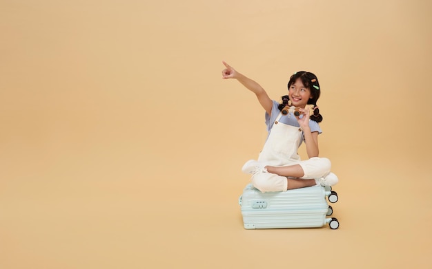 Free photo asian child sitting on suitcase pointing finger at copy space and look through binoculars