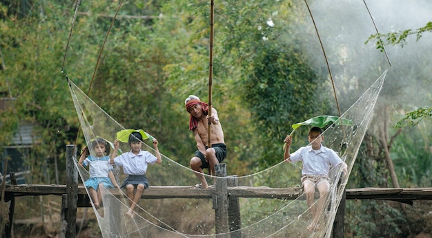 Asian children enjoy to cheer up grandfather or senior fisherman use square dip net to catching fish in canal, Three sibling cheerful on wooden bridge after school, copy space, rural lifestyle concept
