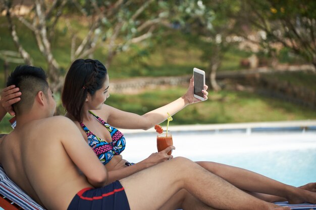 Asian couple lying on loungers by swimming pool and taking selfies on smartphone
