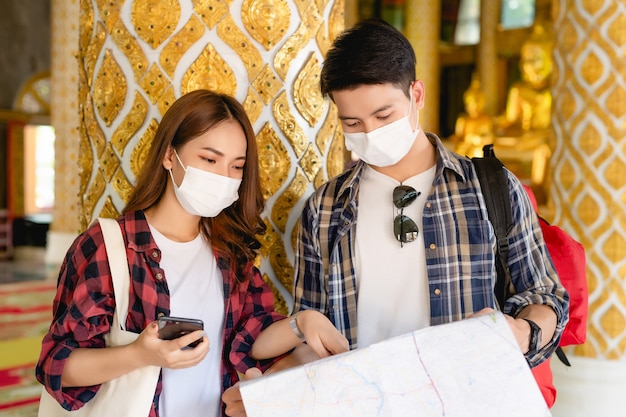 Coppia asiatica di turisti zaino in spalla in piedi nel bellissimo tempio tailandese, bella donna che tiene in mano uno smartphone e un bell'uomo che controlla la mappa cartacea durante il viaggio in vacanza