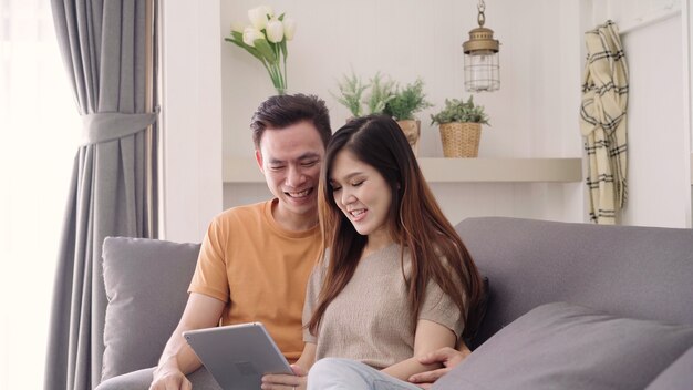 アジアのカップルが自宅の居間でインターネットでのオンラインショッピングのためのタブレットを使用して、甘いカップルを楽しむ