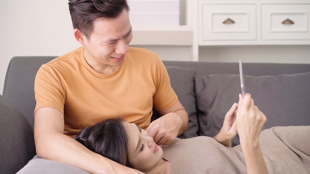 アジアのカップルが自宅の居間でインターネットでのオンラインショッピングのためのタブレットを使用して、甘いカップルを楽しむ