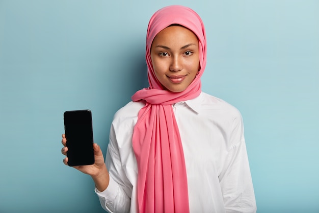 Free photo asian dark skinned female in pink scarf, wears white shirt, holds cell phone with mock up screen for image or text insert, isolated over blue wall. selective focuse. technology, culture, advert