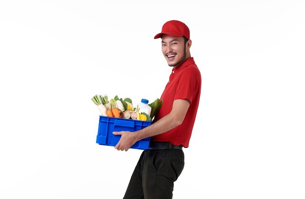 Asian delivery man wearing in red uniform holding fresh food basket isolated over white wall.