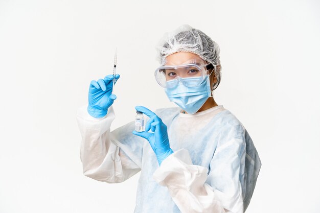 Азиатский врач или медсестра в медицинском защитном снаряжении, уверенно смотрит в камеру, держит шприц и вакцину от covid-19, кампания вакцинации и медицинская концепция