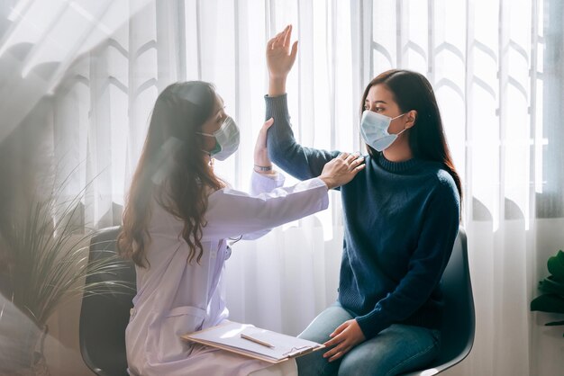 Азиатский врач посещает и осматривает молодую взрослую женщину в больнице с симптомом офисного синдрома. Врач осматривает и консультирует по вопросам здравоохранения в маске для защиты от пандемии covid19