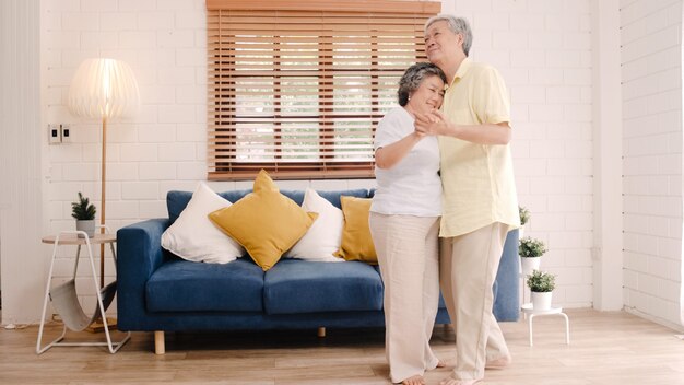 Asian elderly couple dancing together while listen to music in living room at home, sweet couple enjoy love moment while having fun when relaxed at home.