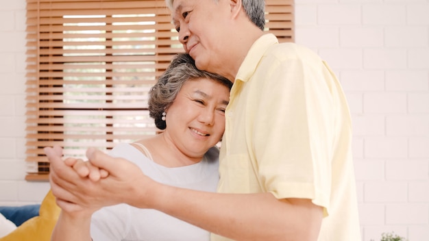 Asian elderly couple dancing together while listen to music in living room at home, sweet couple enjoy love moment while having fun when relaxed at home. 