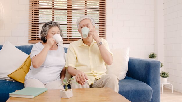 アジアの老夫婦が暖かいコーヒーを飲みながら自宅の居間で話していると、カップルは自宅でリラックスしたときソファの上に横たわっている間愛の瞬間を楽しんでいます。