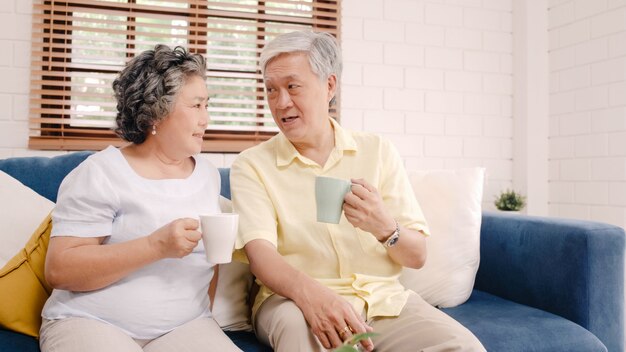 따뜻한 커피를 마시고 집에서 거실에서 함께 이야기하는 아시아 노인 부부는 집에서 편안하게 소파에 누워있는 동안 사랑의 순간을 즐길 수 있습니다.