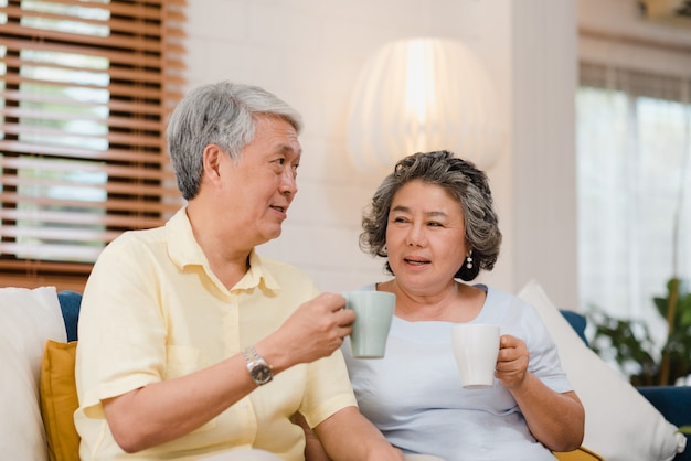 アジアの老夫婦が暖かいコーヒーを飲みながら自宅の居間で話していると、カップルは自宅でリラックスしたときソファの上に横たわっている間愛の瞬間を楽しんでいます。