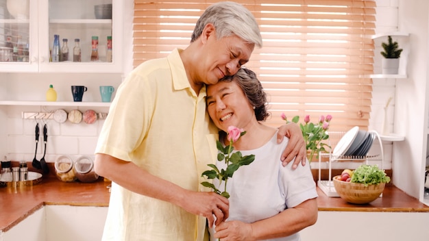 Asian elderly couple feeling happy smiling and holding flower and looking to camera while relax in kitchen at home. Lifestyle Senior family enjoy time at home concept.