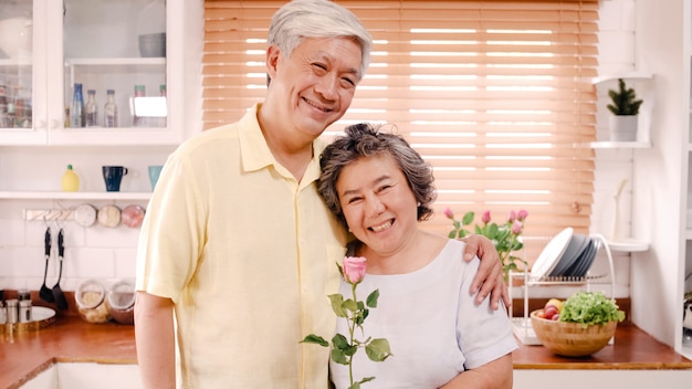 アジアの老夫婦が幸せな笑顔と花を持って自宅のキッチンでリラックスしながらカメラを探しています。