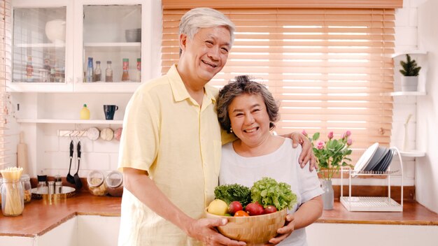 Asian elderly couple feeling happy smiling and holding fruit and looking to camera while relax in kitchen at home. Lifestyle Senior family enjoy time at home concept. 