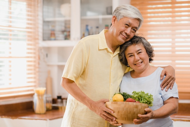 アジアの老夫婦が幸せな笑顔と果物を持って、自宅の台所でリラックスしながらカメラ目線を感じています。