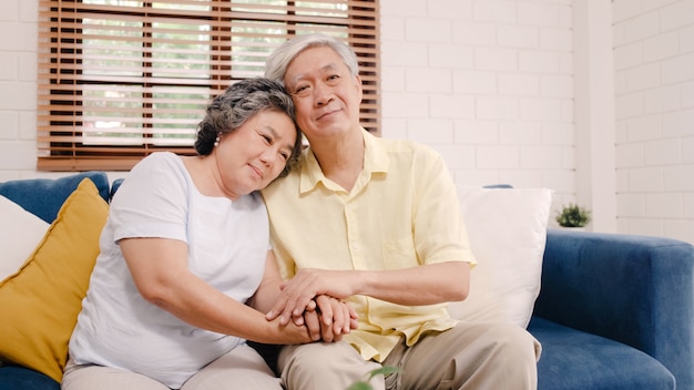 Asian elderly couple holding their hands while taking together in living room, couple feeling happy share and support each other lying on sofa at home. 