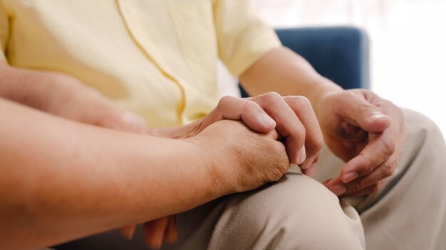 Asian elderly couple holding their hands while taking together in living room, couple feeling happy share and support each other lying on sofa at home. 