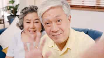 Free photo asian elderly couple using smartphone video conference with grandchild while lying on sofa in living room at home.