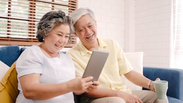 Asian elderly couple using tablet and drinking coffee in living room at home, couple enjoy love moment while lying on sofa when relaxed at home. 