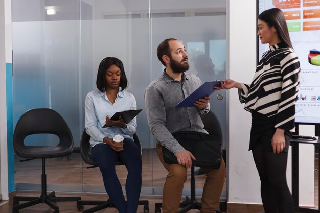 Asian entrepreneur giving cv resume to man recruiter asking about work experience during interview meeting in startup company office. Diverse candidates waiting for career opportunity in lobby area