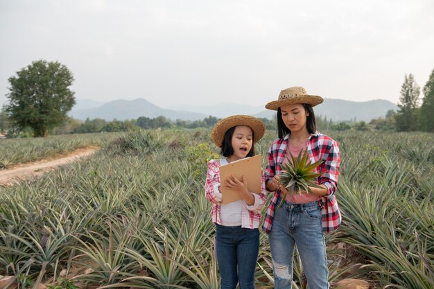 アジアの農家は、母と娘に農場でのパイナップルの成長を見てもらい、データをクリップボードの農家のチェックリストに保存します。