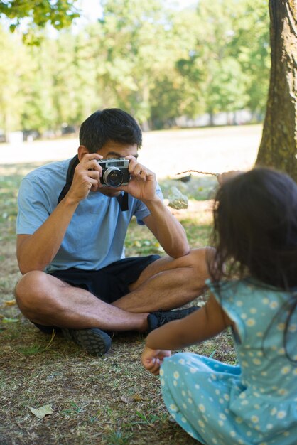 아시아 아버지 여름 공원에서 좋은 딸의 사진을 찍고. 카메라를 들고 있는 젊은 남자와 잔디밭에 앉아 있는 어린 소녀가 아빠를 바라보며 포즈를 취하고 있습니다. 어린이 활동적인 여가와 행복한 어린 시절 개념