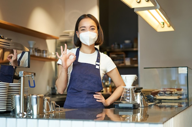 Foto gratuita la ragazza barista asiatica in maschera medica mostra che il segno ok funziona nel bar dietro il bancone prepara il caffè