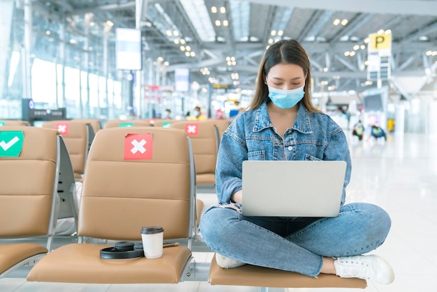 아시아 여성 캐주얼 천은 바이러스 보호 얼굴 마스크를 착용하고 사회적 좌석 거리를 둔 새로운 일반 생활 방식을 통해 공항 터미널 안전 여행 개념에서 손으로 일하는 노트북을 즐깁니다.