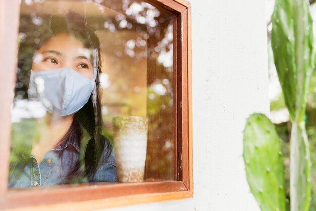 Азиатская женская повседневная рубашка носит маску для лица защита от вирусов новый нормальный образ жизни сидит расслабляется рядом с оконным стеклом с отражением сада за пределами концепции здоровья и идей