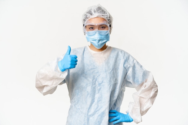 Asian female doctor or nurse wearing personal protective equipment and rubber gloves, medical face mask, showing thumbs up, vaccination and covid-19 campaign concept