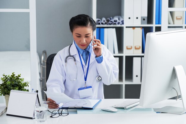 Азиатский женский доктор сидя на столе в офисе и вызывая на мобильном телефоне