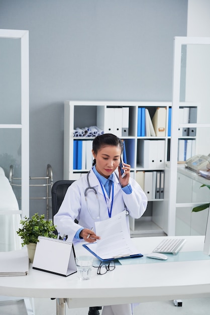 Foto gratuita medico femminile asiatico che parla sul telefono cellulare in ufficio e che esamina le annotazioni