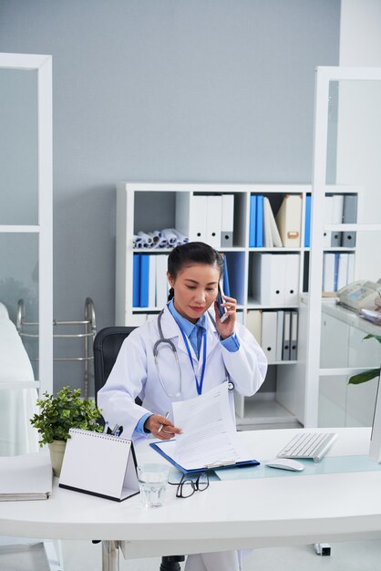 アジアの女性医師がオフィスで携帯電話で話していると記録を見て