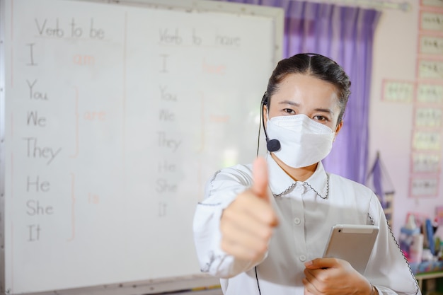 Asian female english teacher wearing a mask teaching students to study online on a computer screen by using an online video conferencing system for education