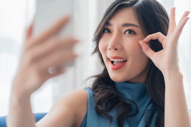 아시아 여성 제스처 재미있는 비디오 쾌활한 행복 대화로 친구에게 전화하는 아시아 여성 얼굴 표정 손 제스처는 소파에서 괜찮습니다.